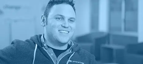 Smiling man wearing a dark zip-up hoodie and plaid shirt, sitting indoors.