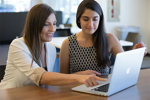 Two women working together and looking at a laptop on a wooden table in a modern office.
