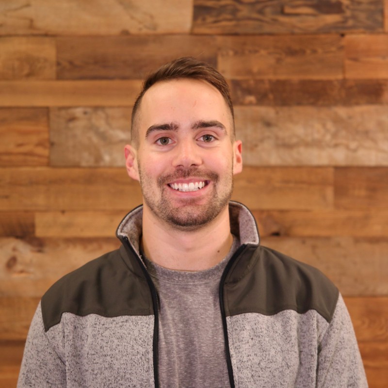 Smiling young man with short brown hair and beard wearing a gray and black zip-up jacket in front of a wooden wall.