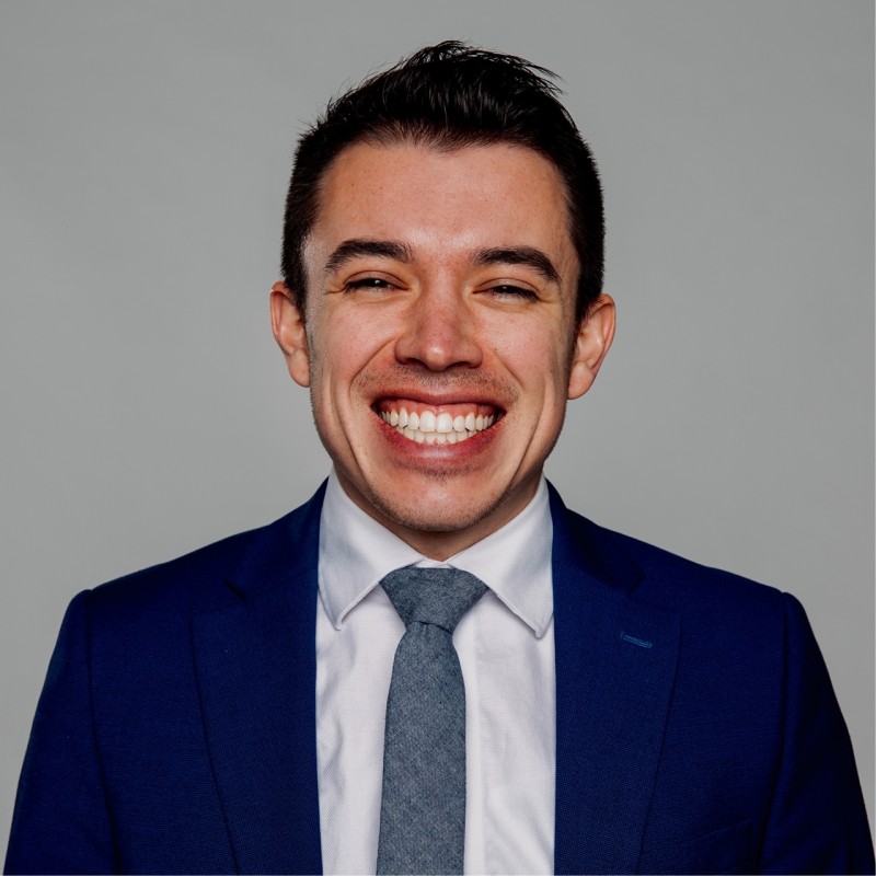 Smiling man in a blue suit, white shirt, and gray tie against a gray background.