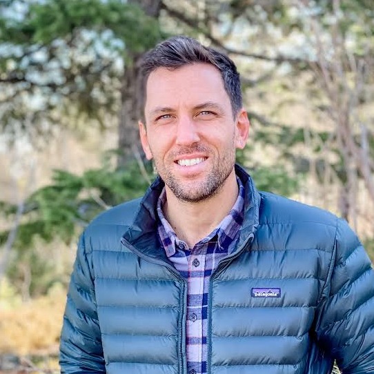 Smiling man wearing a blue puffer jacket and plaid shirt standing outdoors with trees in the background.