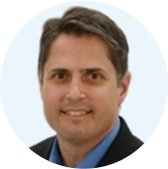 Portrait of a smiling man with short brown hair wearing a dark blazer and blue shirt, against a light blue background.