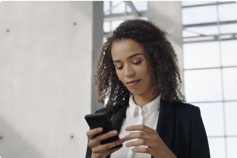 Woman with curly hair wearing a black blazer and white shirt looking at her smartphone in a modern office space.