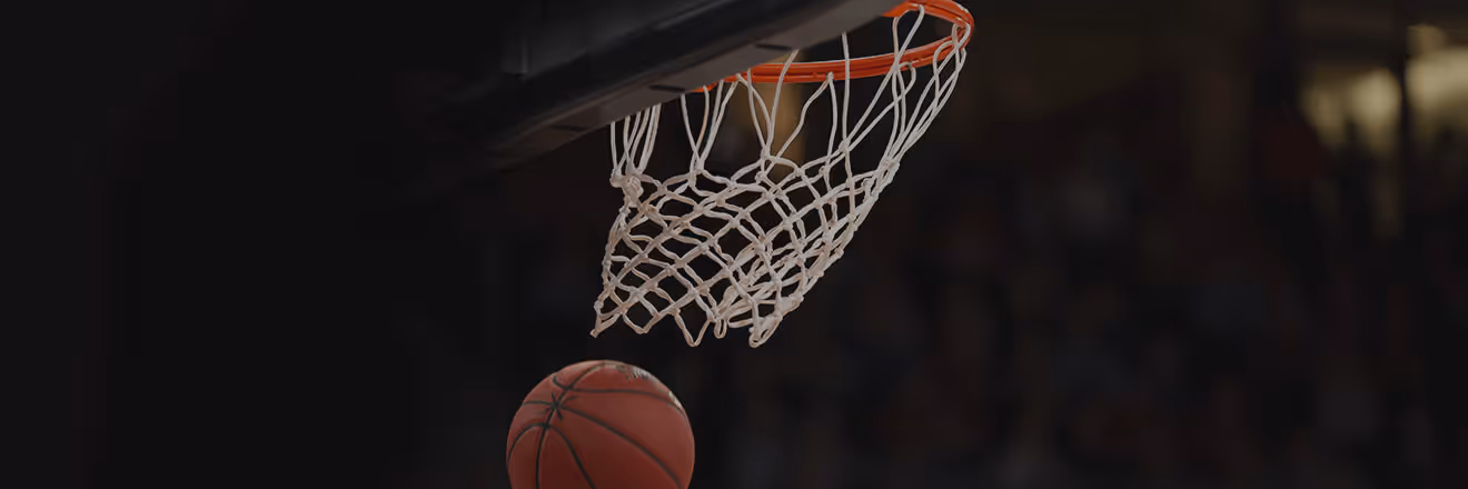 Basketball approaching an orange hoop with white net during a game in a dark arena.