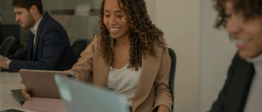 Smiling woman with curly hair using a tablet at a table with two colleagues in a professional setting.