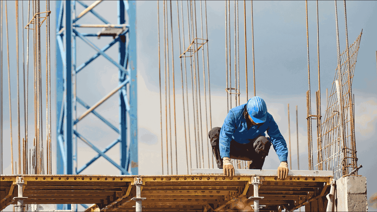 Construction worker wearing a blue helmet and jacket working on a building framework with metal rods and scaffolding.