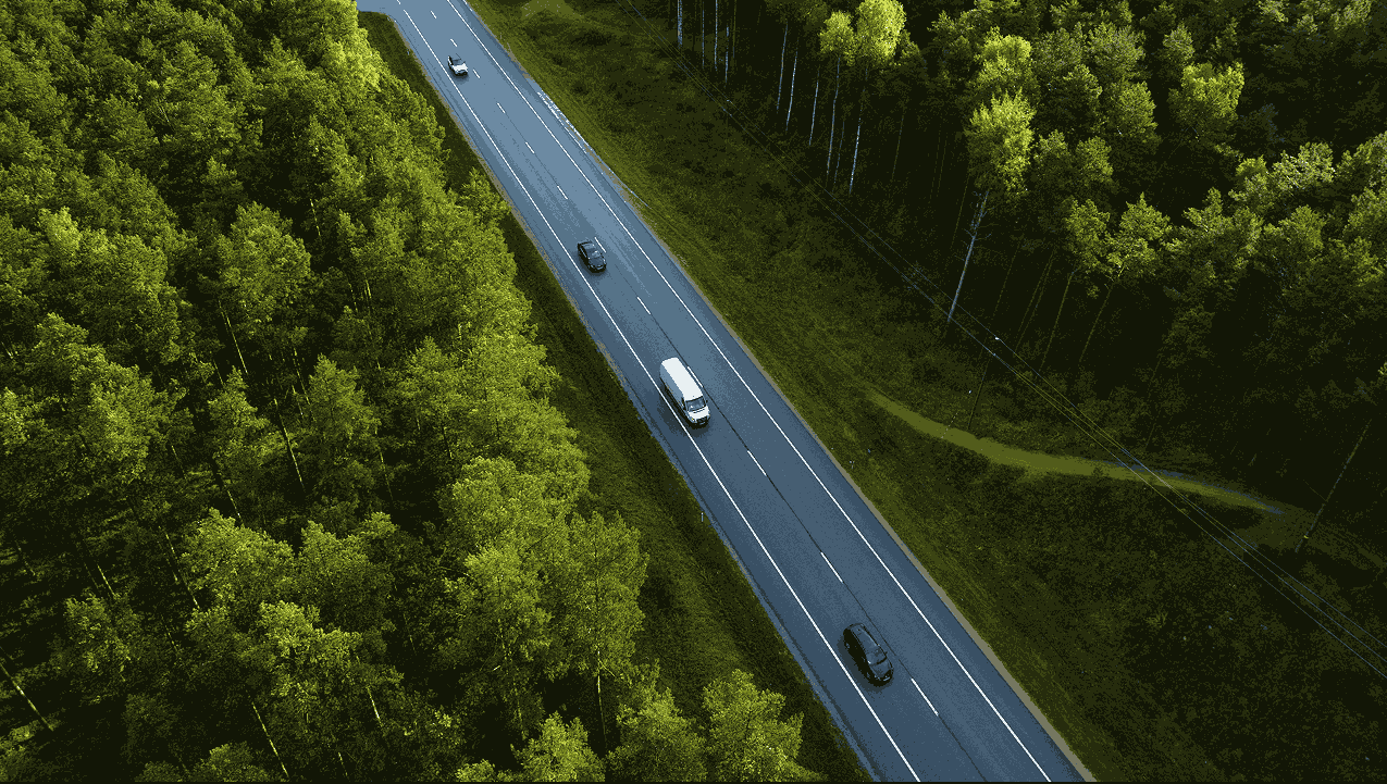 Aerial view of a straight two-lane road running through a green forest with three vehicles driving on it.