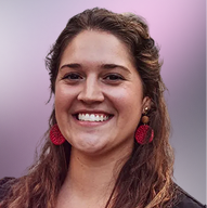 Smiling woman with long curly hair wearing red round earrings and a purple top against a soft purple background.