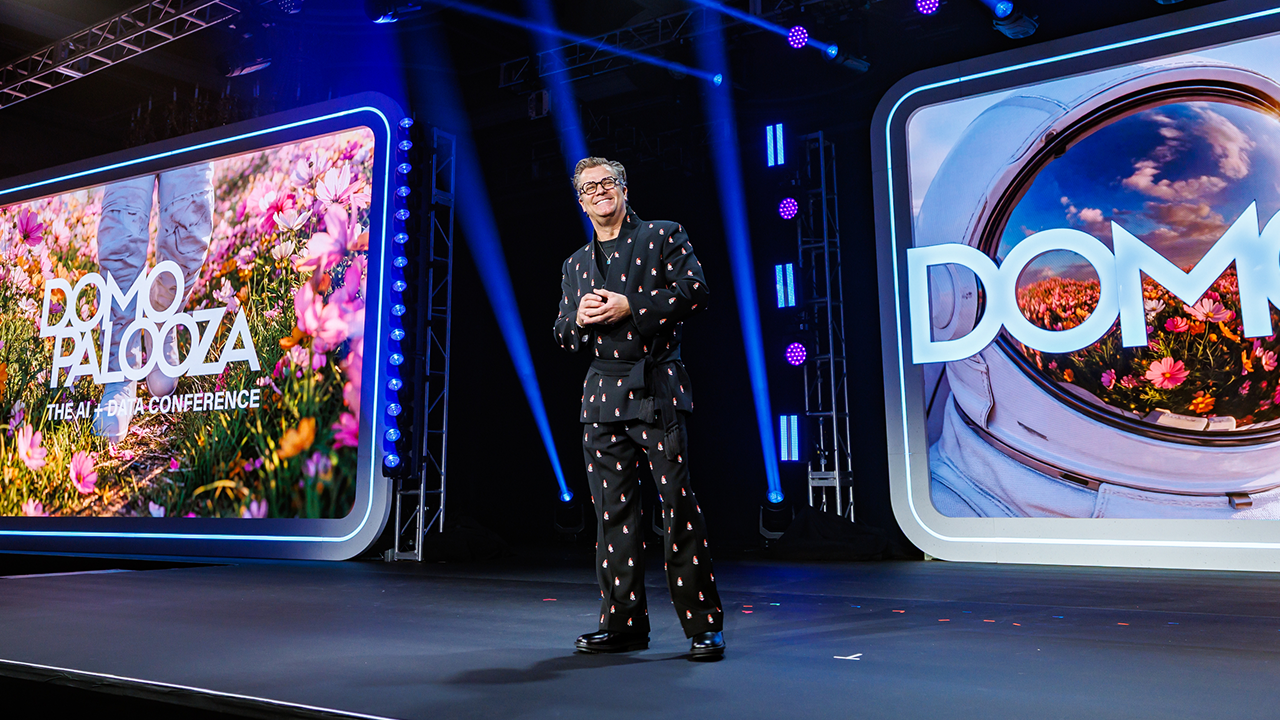 A man in a patterned black suit stands on stage with two large screens behind him displaying 'DOMO PALOOZA THE AI + DATA CONFERENCE' and floral and astronaut helmet visuals.