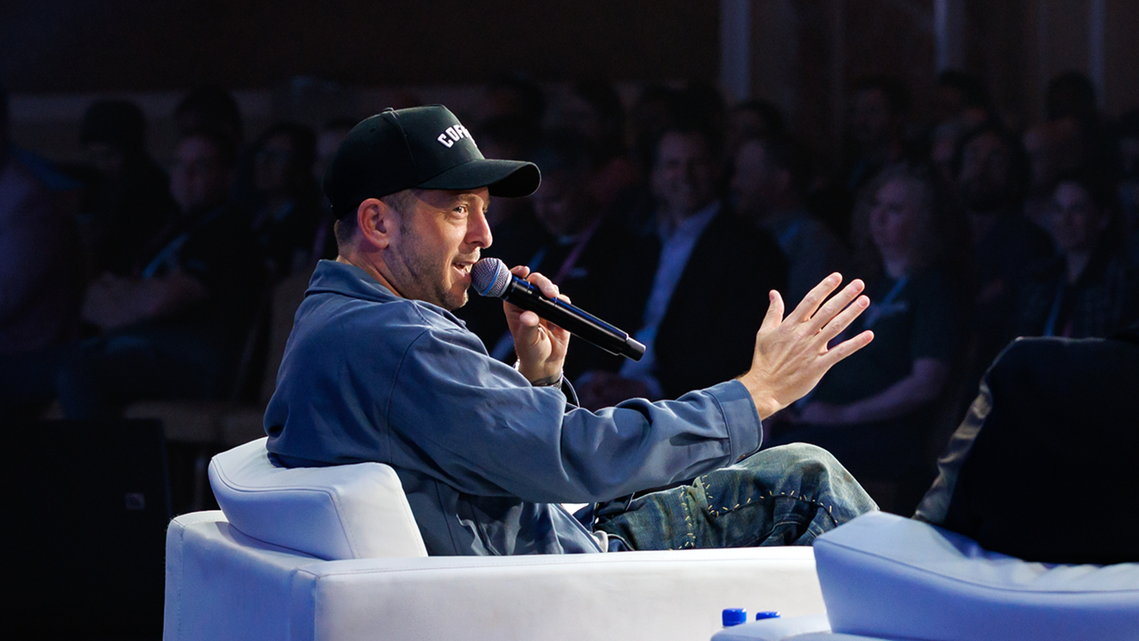 Man in casual clothes and a black cap speaking into a microphone while seated on a white armchair in front of an audience.