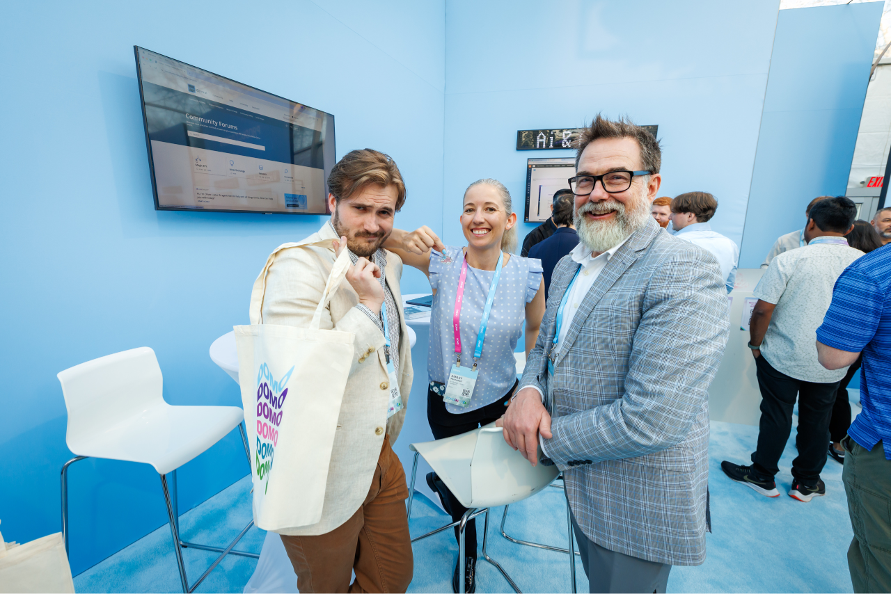 Three people posing and smiling at a tech event booth, one man holding a tote bag with repeated 'DOMO' text.
