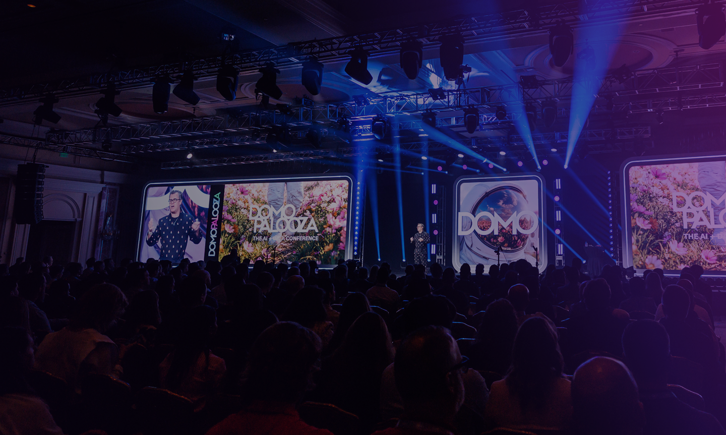 Speaker presenting on stage at Domo Palooza conference with audience facing large digital screens and blue stage lighting.