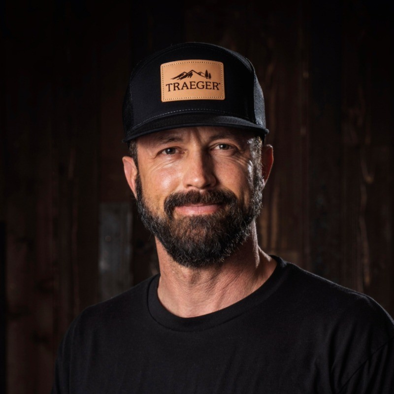 Bearded man wearing a black Traeger cap and black shirt smiling against a dark wooden background.