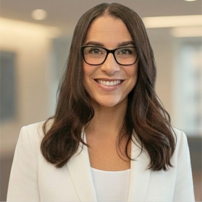 Smiling woman with dark hair wearing glasses and a white blazer in a blurred office background.