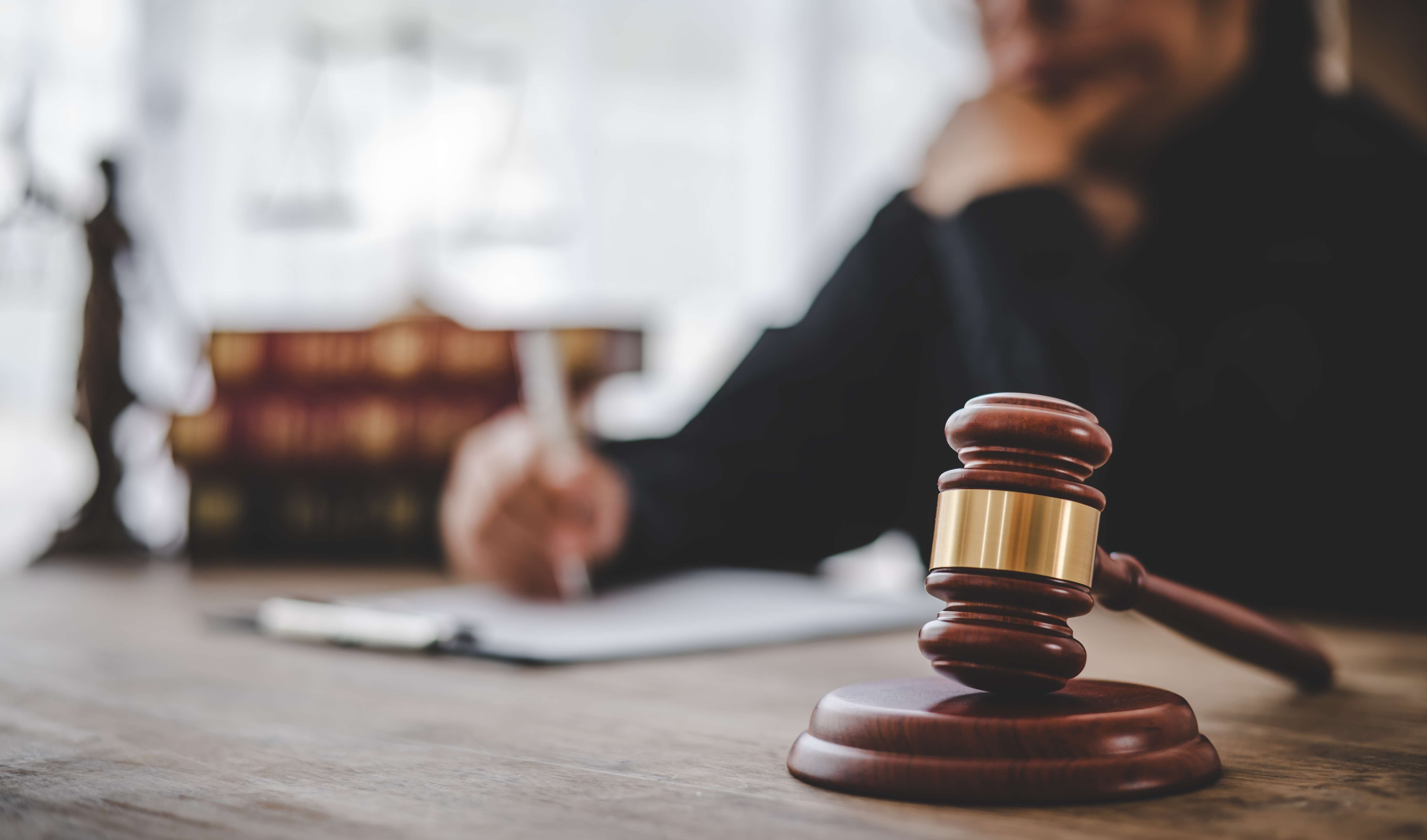 Wooden judge’s gavel on desk with person writing on paper in blurred background.