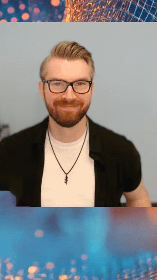 Smiling man with glasses, beard, and pendant necklace wearing a black shirt over a white tee.