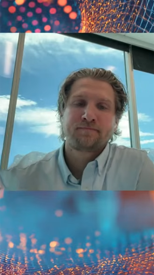Man with light beard and wavy hair wearing a light blue shirt in front of a window with a blue sky view.