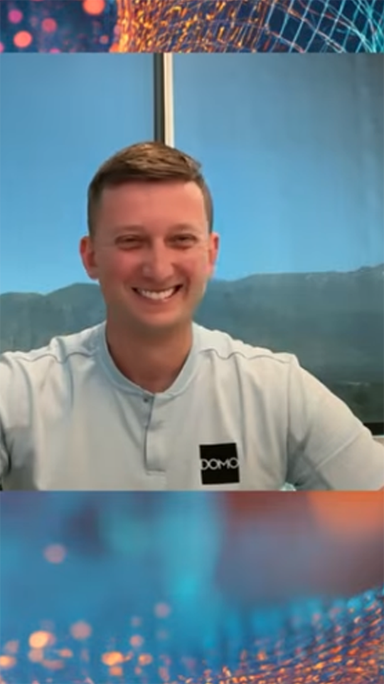 Smiling man in a light blue shirt with DOMO logo, sitting indoors with a mountainous view behind him.