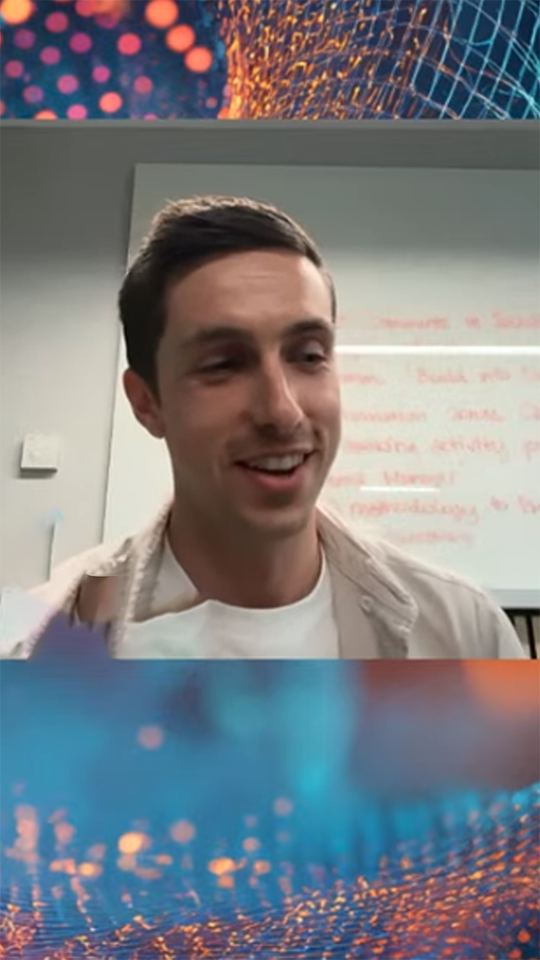 Smiling young man with short dark hair in front of a whiteboard with blurred writing.