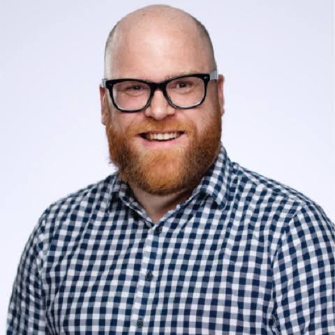 Smiling bald man with red beard and black glasses wearing a checkered shirt on a white background.