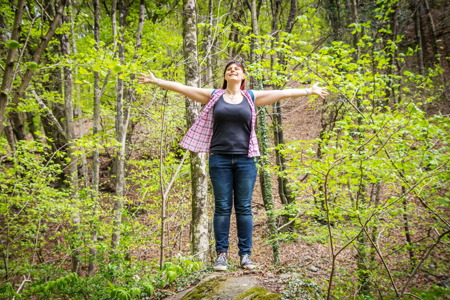 Smiling woman standing on a rock in the forest with arms wide open, symbolizing freedom, healing, and a fresh start in addiction recovery