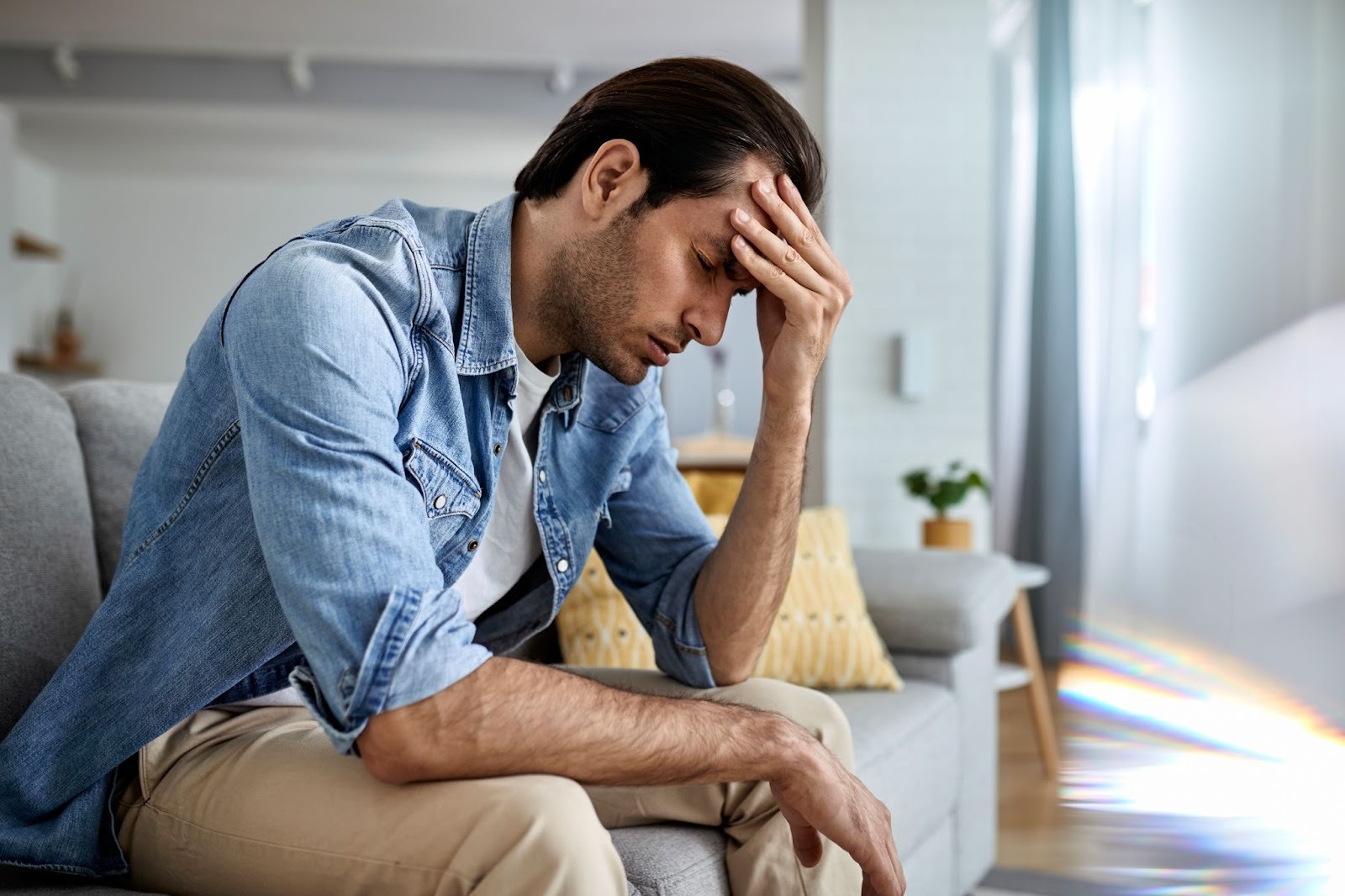 A man sits on a couch with his head in his hand, appearing fatigued and anxious, representing the symptoms of alcohol withdrawal