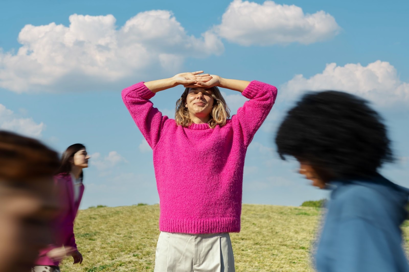 A woman wearing a pink sweater stands amidst a vibrant green field, surrounded by nature on a sunny day
