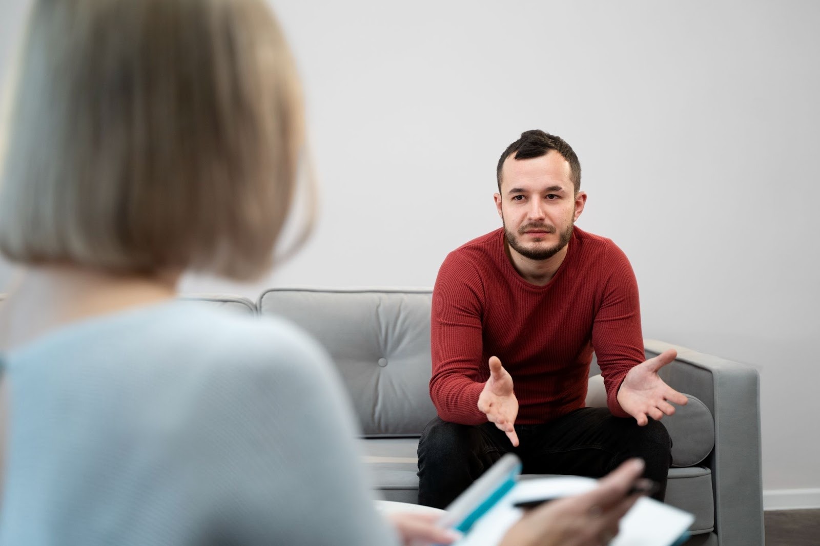 A man in a red sweater sits on a couch, gesturing with his hands while talking to a woman holding a clipboard