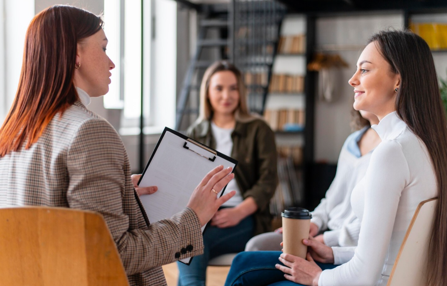 A medium shot of a group therapy session, with two women in the foreground talking to each other