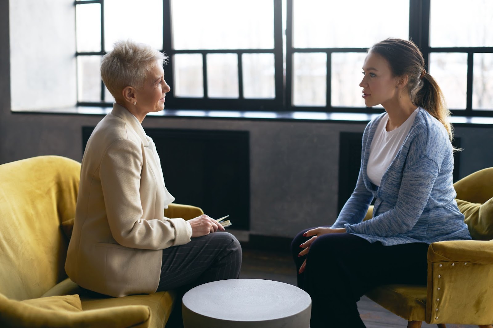 Two women sit facing each other in a well-lit room, one holding a notebook, reflecting the focused, professional guidance central to licensed clinicians in addiction treatment