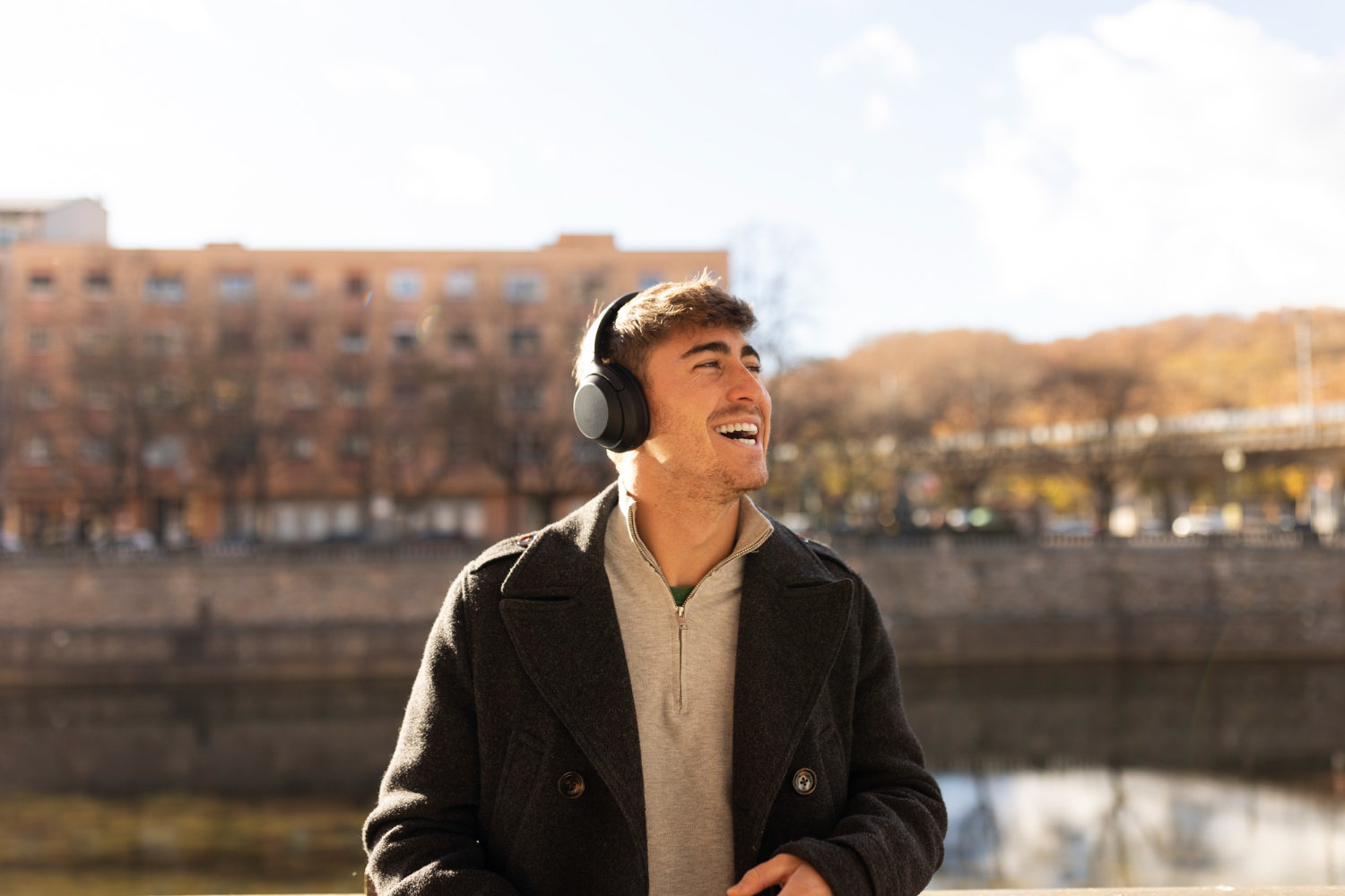 Young man wearing headphones and a jacket, smiling while standing near a river on a sunny day, representing calm, reflection, and personal well-being