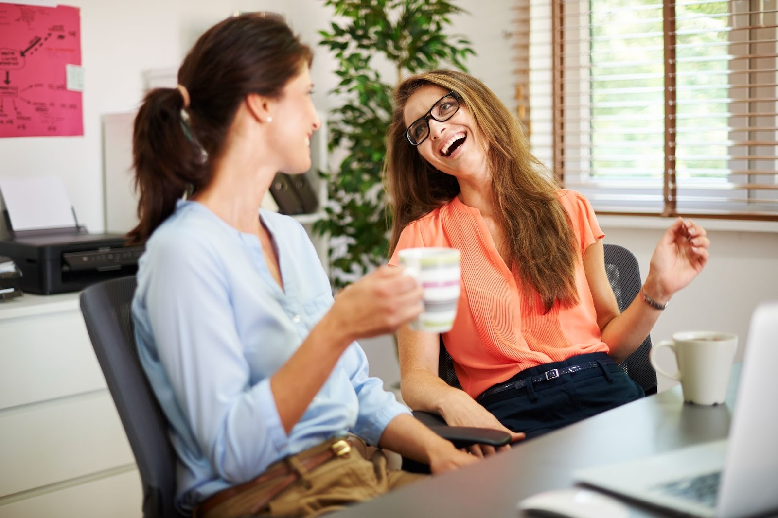 Two women sit in an office, smiling and laughing with coffee mugs, symbolizing support and connection while overcoming high-functioning addiction
