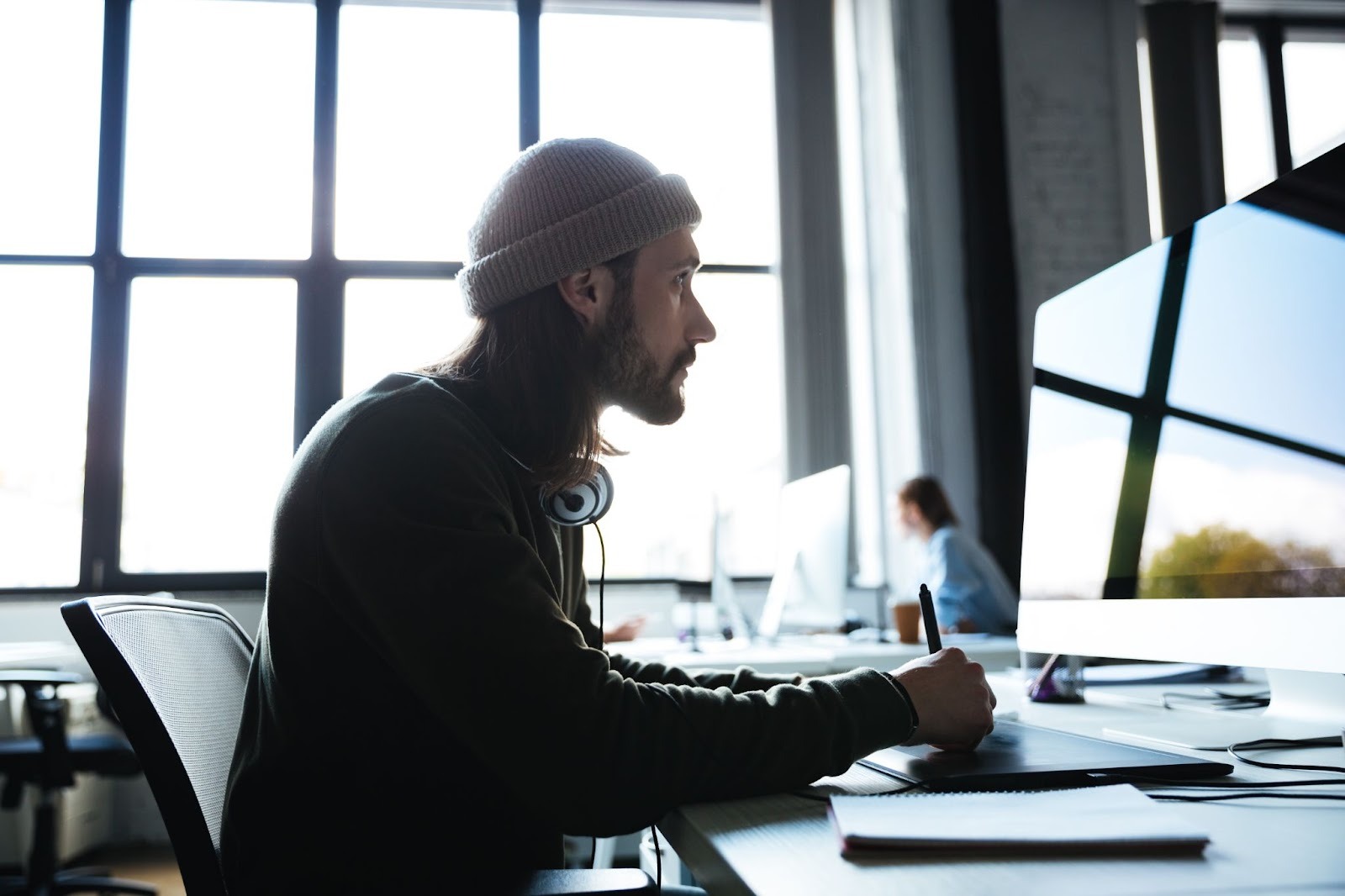 A person with long hair, wearing a beanie, works intently at a computer in a modern office, illustrating someone hiding a high-functioning addiction while appearing focused and productive