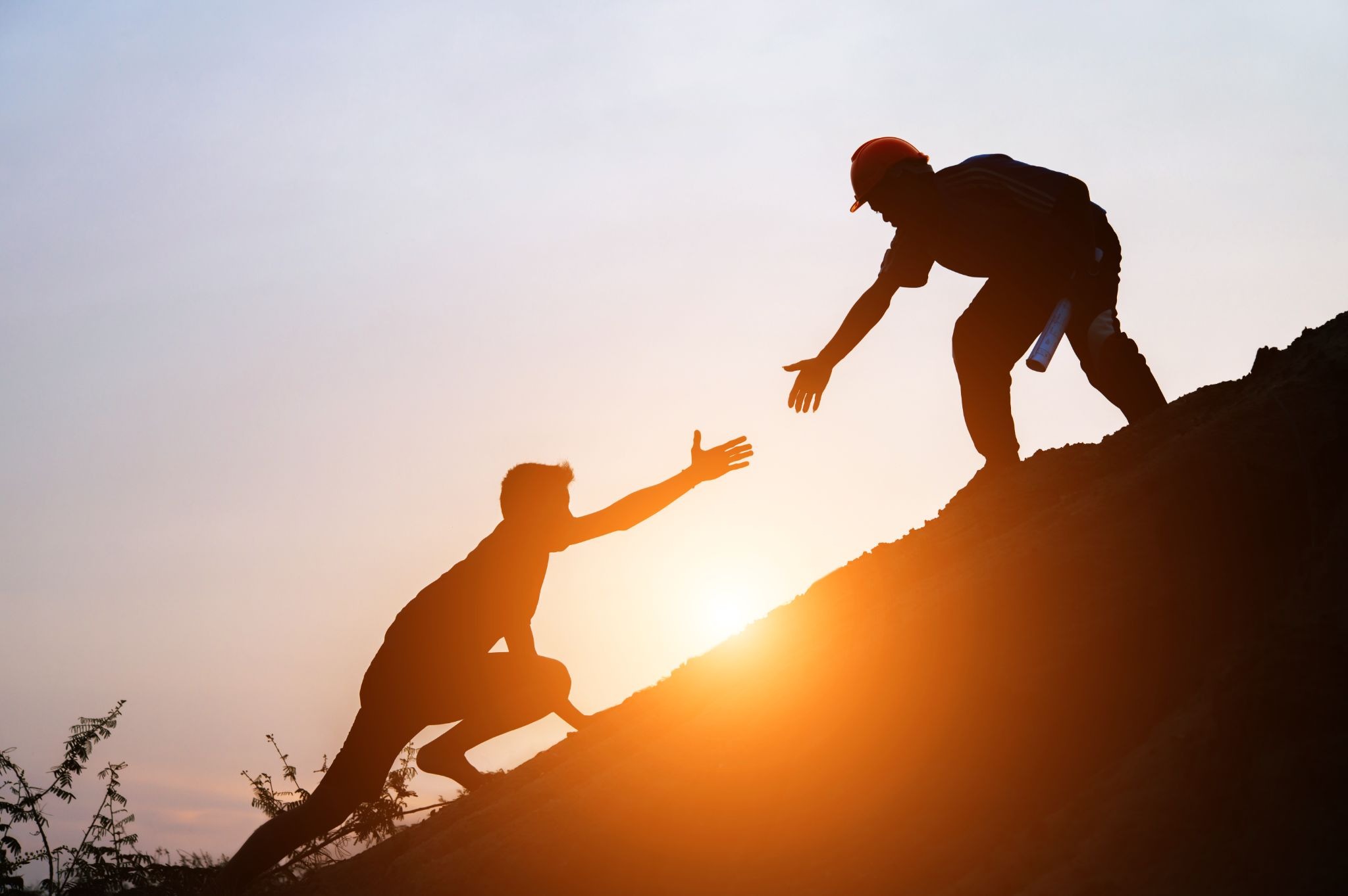 Silhouette of one person helping another climb a steep hill at sunset, symbolizing mentorship, guidance, and recovery support through challenging moments