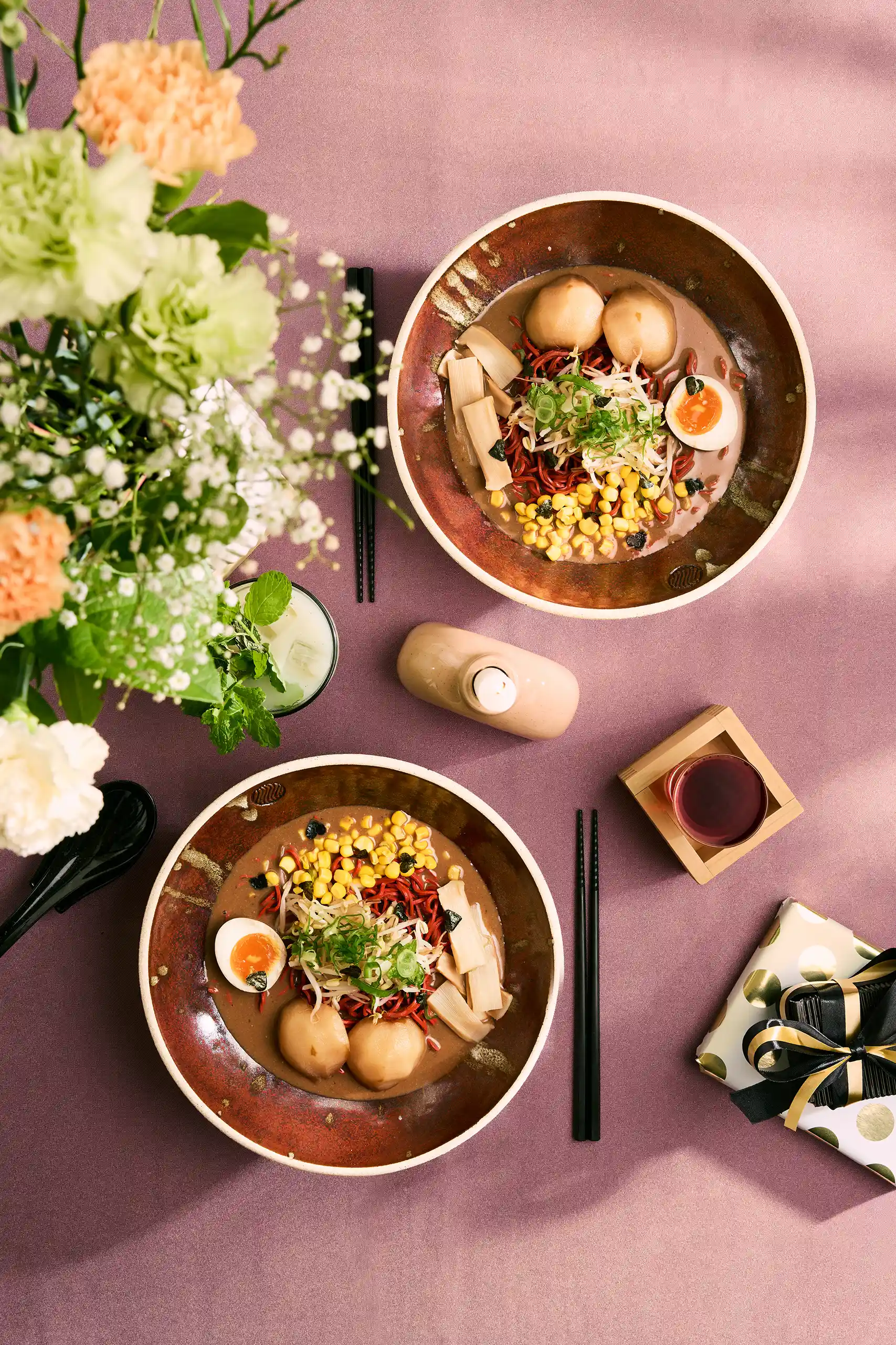 Two bowls of ramen with corn, soft-boiled eggs, mushrooms, and green onions on a purple tablecloth with chopsticks, a glass of drink, flowers, and a wrapped gift box.