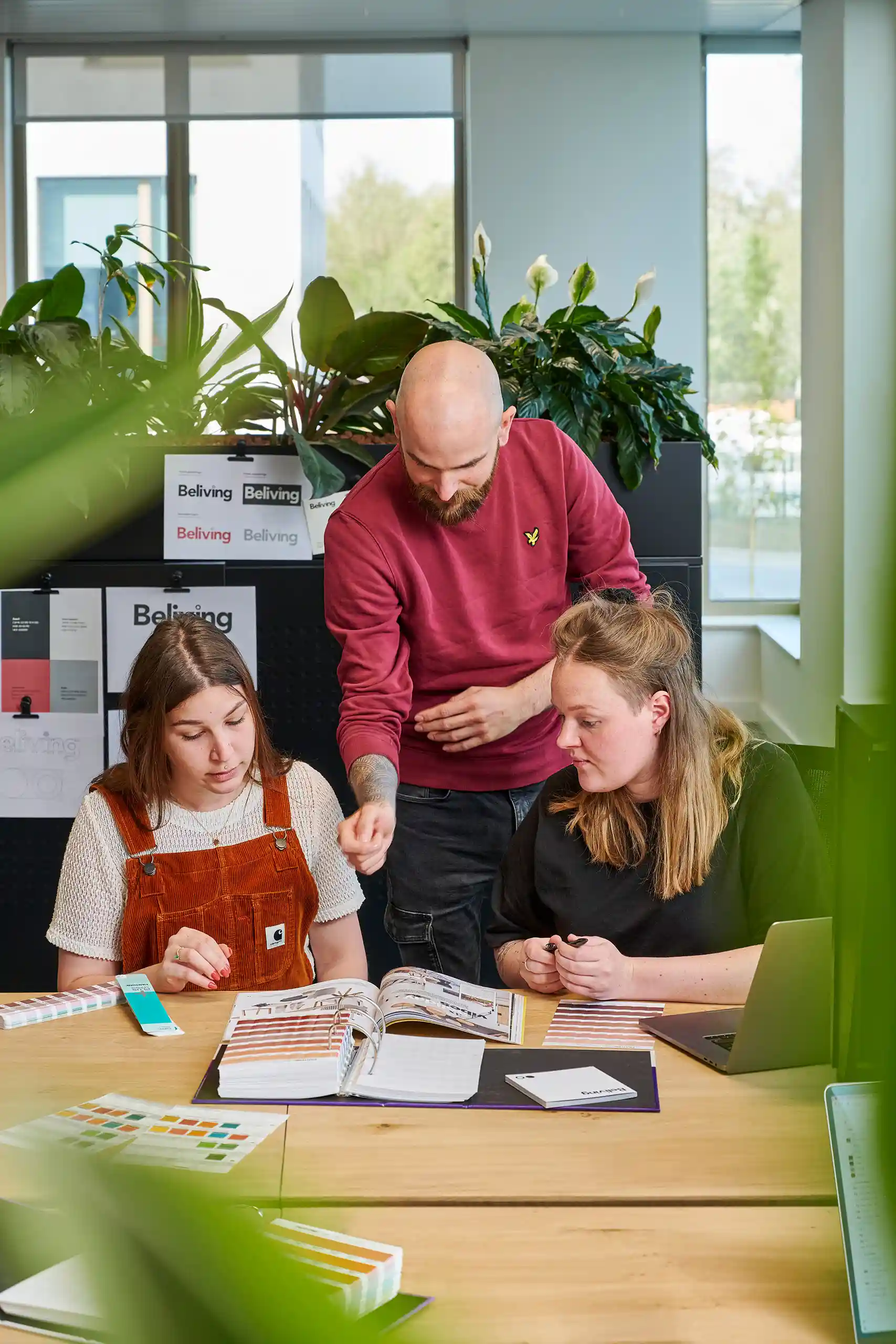 Three people collaborating at a wooden desk with color swatches, open notebooks, and a laptop in a bright office with plants.