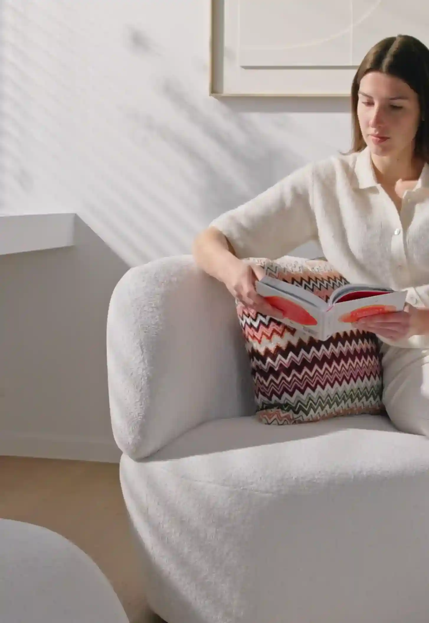 Woman in light-colored outfit reading a book while sitting on a white textured armchair with a colorful zigzag-patterned pillow.