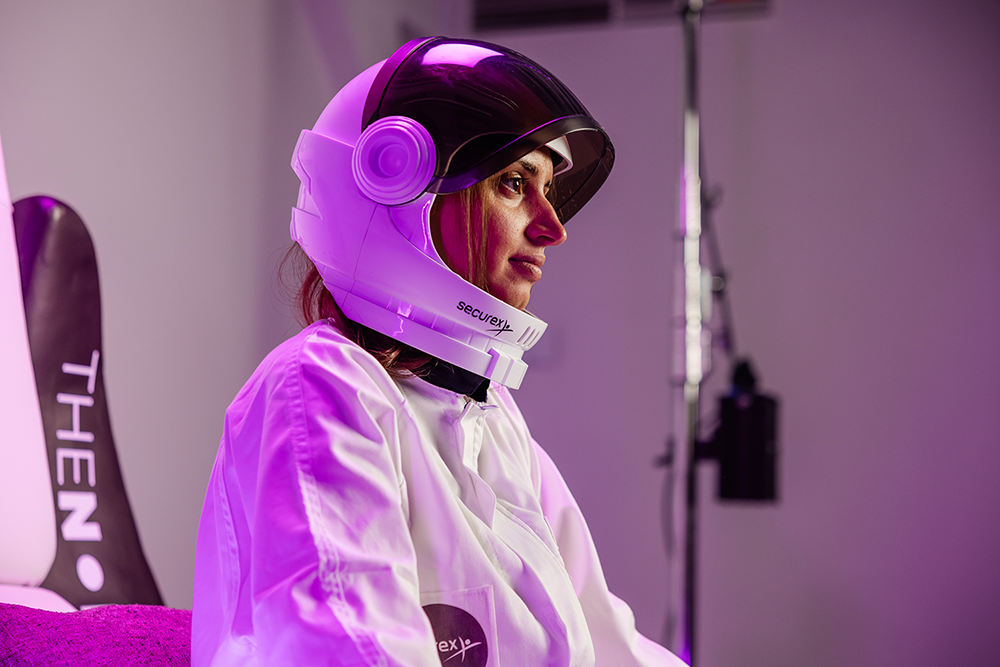 Woman in a white lab coat wearing a futuristic helmet with a dark visor, sitting indoors under purple lighting.
