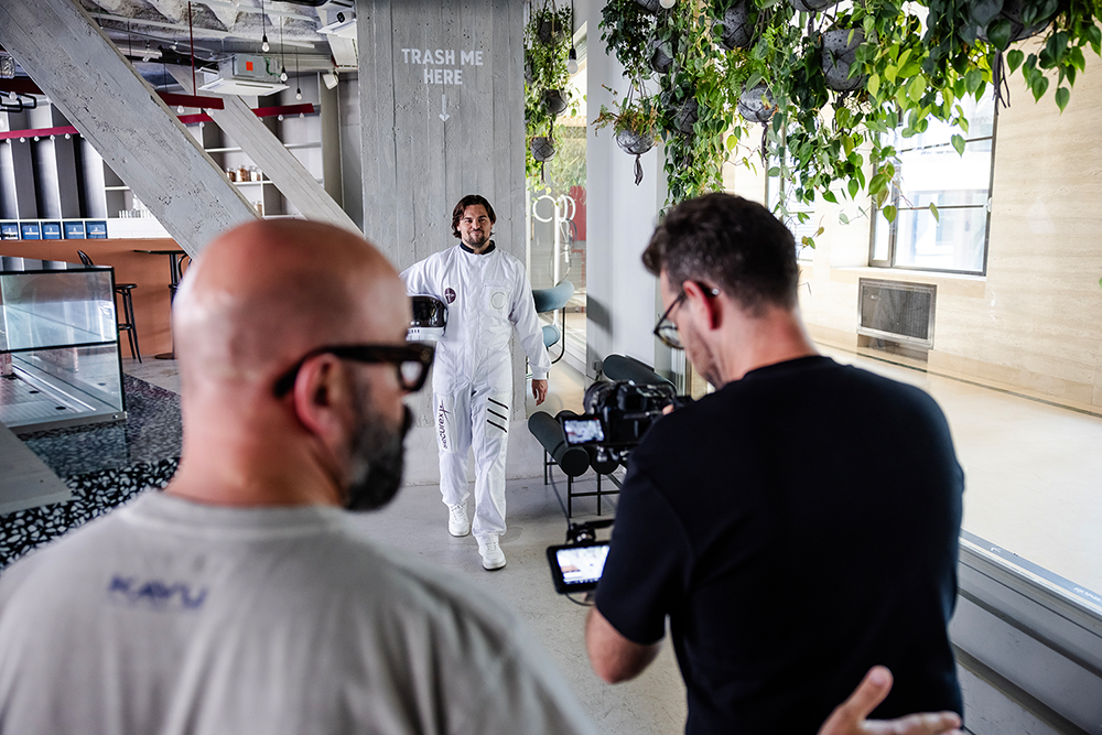 Man in white jumpsuit walking towards two men, one filming with a camera, in a modern indoor setting with plants hanging from the ceiling and a concrete pillar labeled 'TRASH ME HERE'.