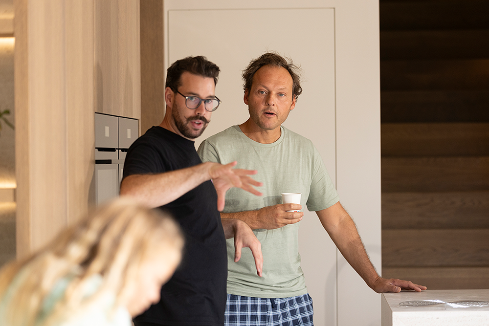 Two men talking indoors, one wearing glasses and a black shirt gesturing with his hand, the other in a green shirt holding a cup.