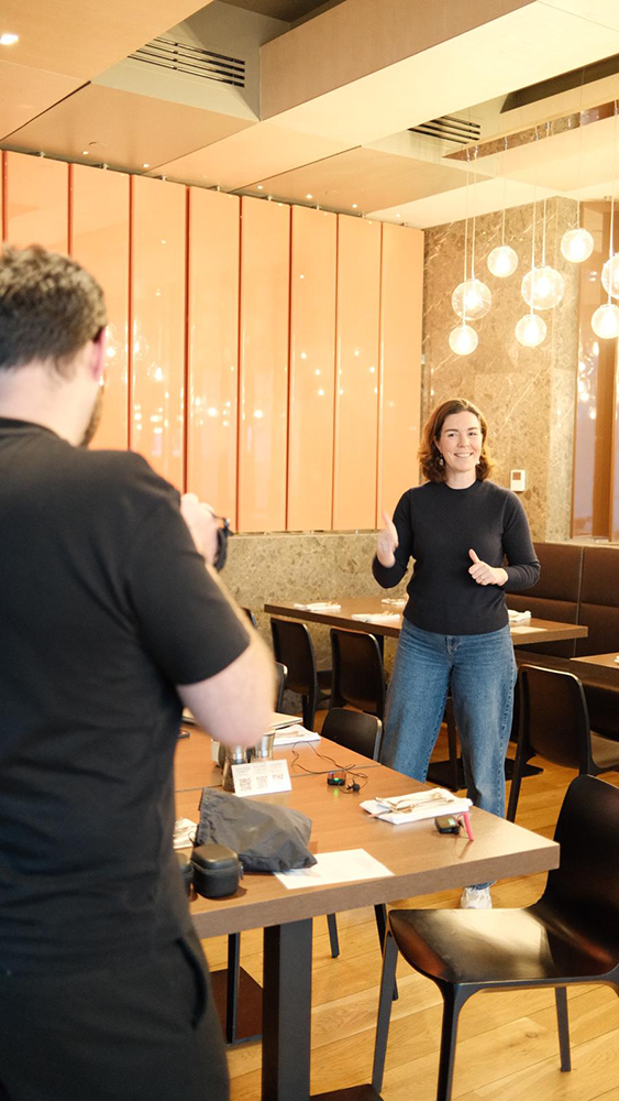Woman in black sweater and jeans giving thumbs up while a man in black takes a photo of her in a modern restaurant.