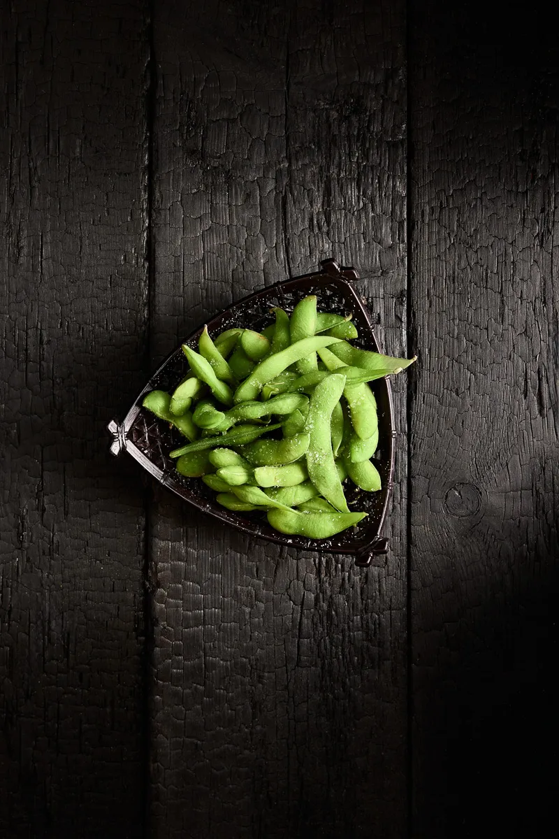 Bowl of green edamame pods sprinkled with salt on a dark textured wooden surface.