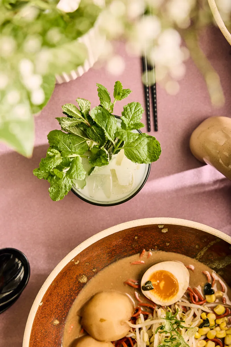 Top view of a bowl of ramen with soft-boiled egg, bean sprouts, corn, and green onions next to a glass of iced mint drink on a pink tablecloth.