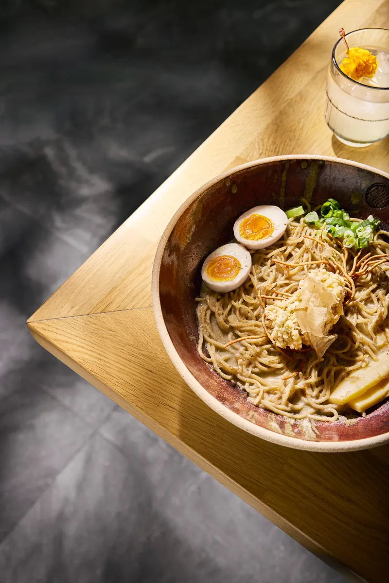 Bowl of ramen with halved soft-boiled egg, noodles, green onions, and tempura on a wooden table next to a glass with a yellow flower garnish.