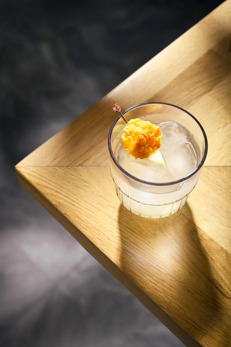 Glass of iced drink garnished with a bright yellow flower on a wooden table corner.