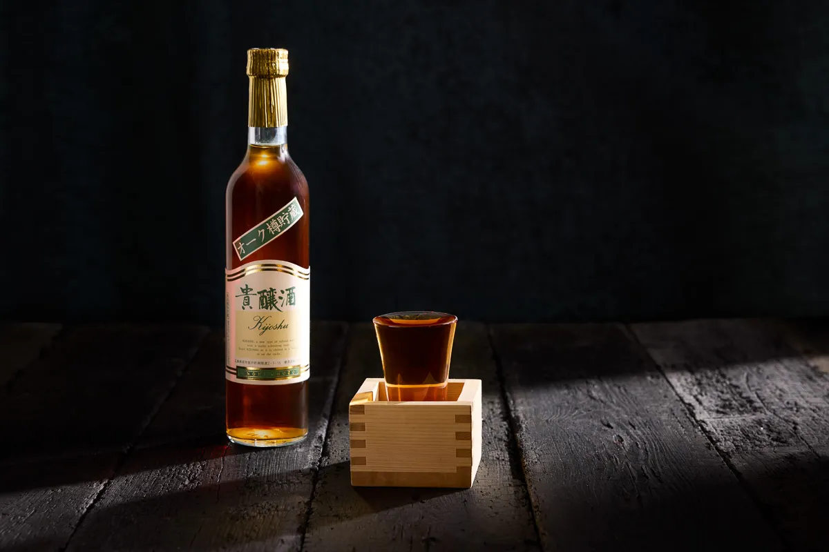 Bottle of Kijoshu sake with a filled glass inside a wooden masu box on a dark wooden surface.