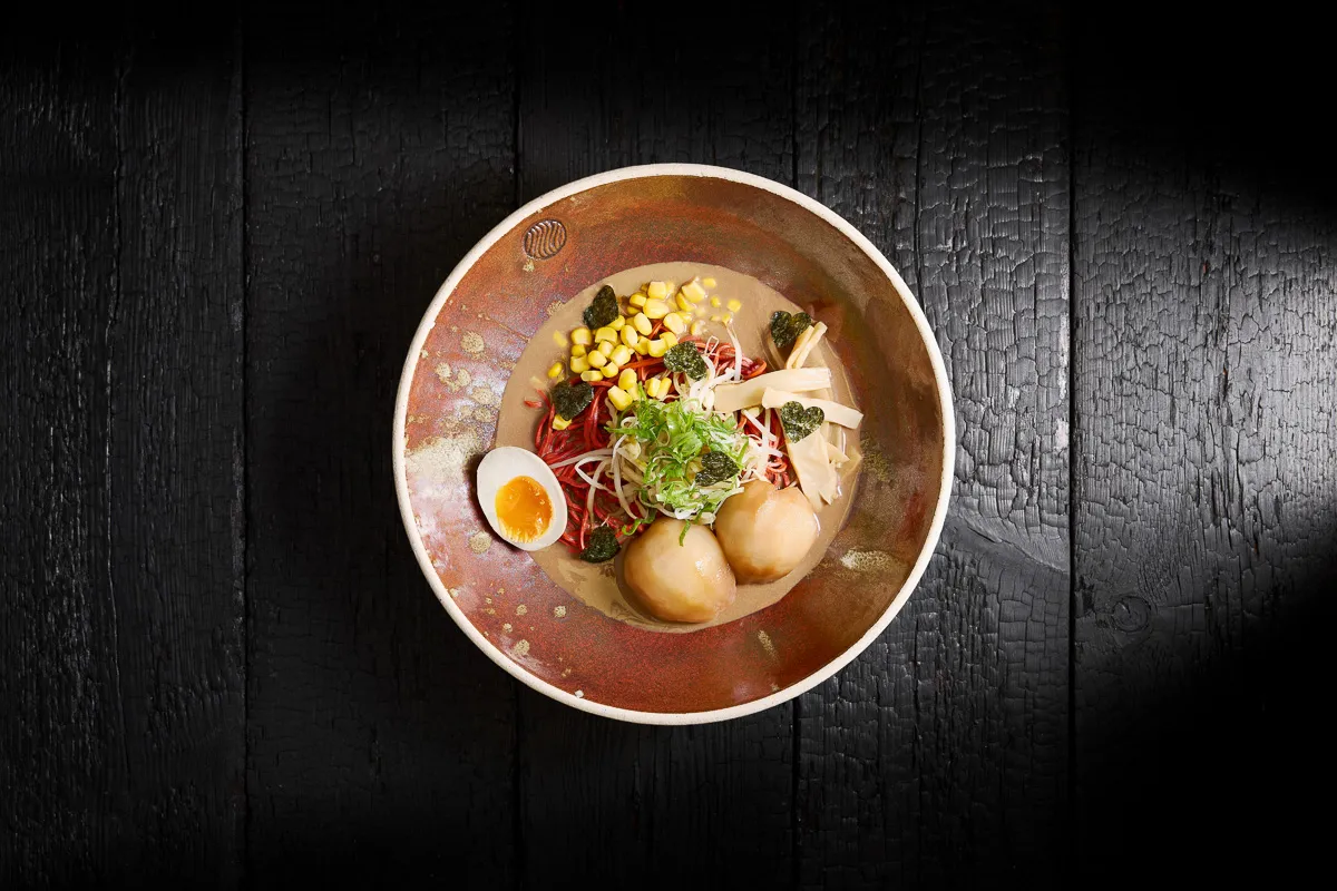 Bowl of ramen with red noodles, corn, bamboo shoots, soft boiled egg, green onions, two dumplings, and heart-shaped seaweed pieces on a dark wooden surface.