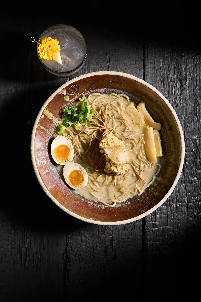 Bowl of ramen with soft-boiled eggs, green onions, bamboo shoots, and crispy garnish in creamy broth next to a cocktail with ice and a yellow flower garnish.