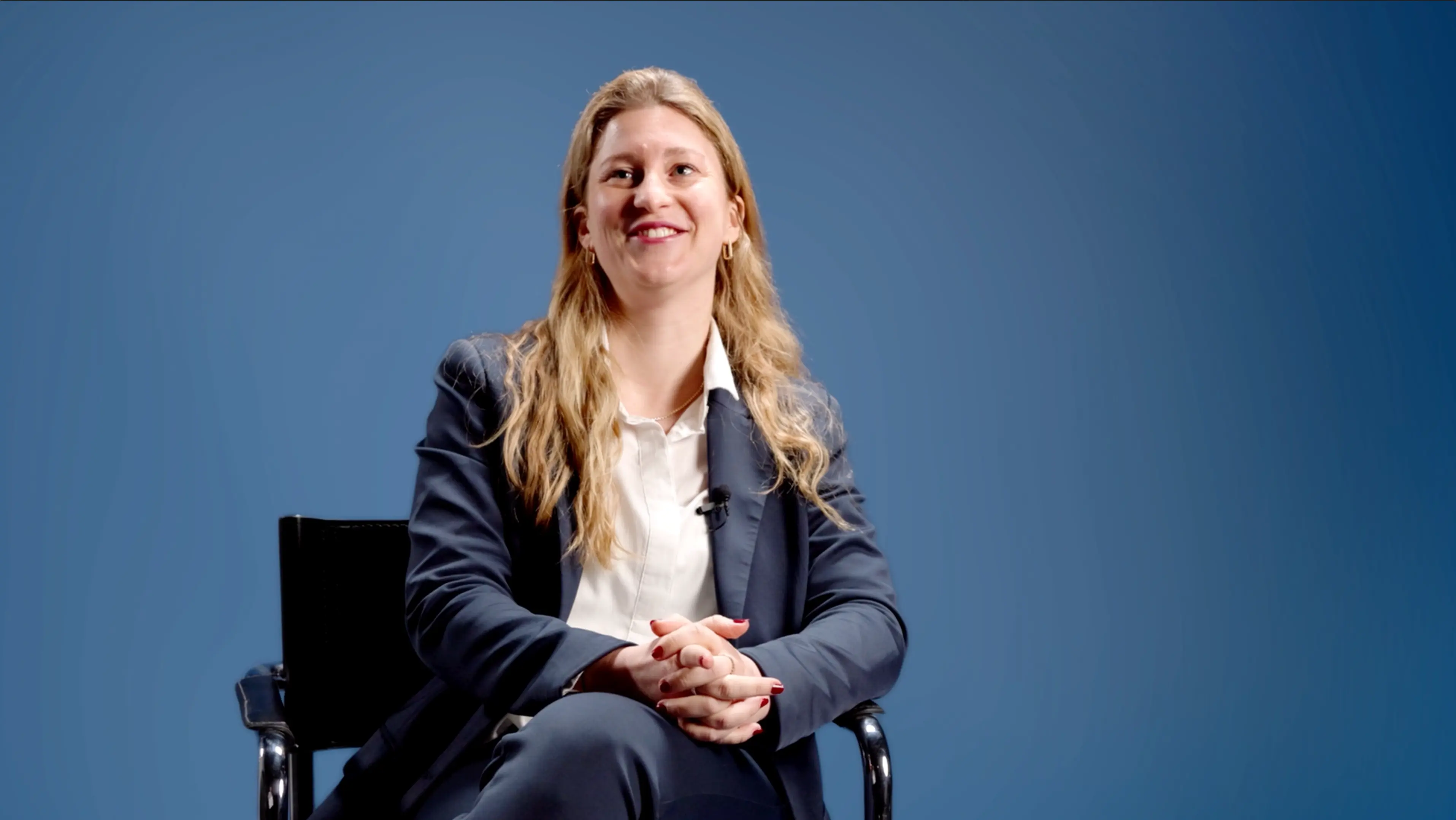Smiling woman with long blonde hair wearing a navy blue suit sitting on a chair against a blue background.