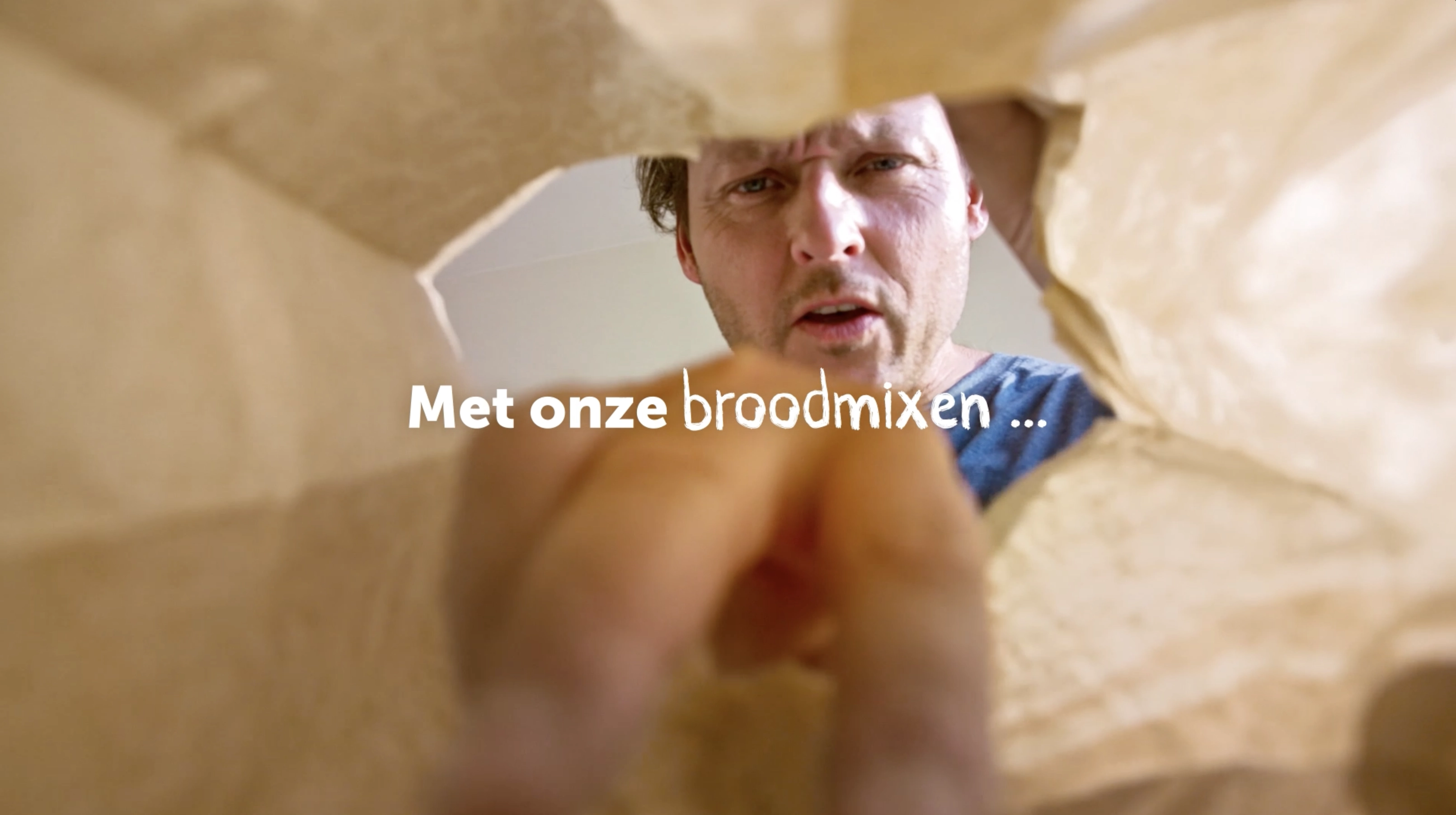 View from inside a paper bag looking up at a man reaching in, with text overlay in Dutch saying 'Met onze broodmixen ...'.